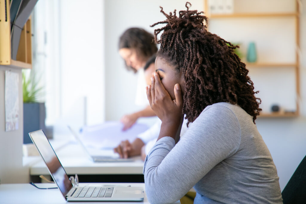 an African American woman working at a computer in the office with her hands rubbing her face